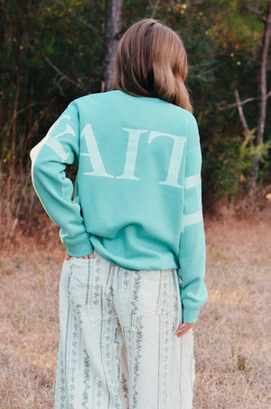 Teal sweatshirt with a white "Faith" back print on a woman standing in a field near trees.