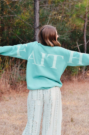 Woman in teal sweatshirt with "FAITH" in white lettering on the back, outdoors.