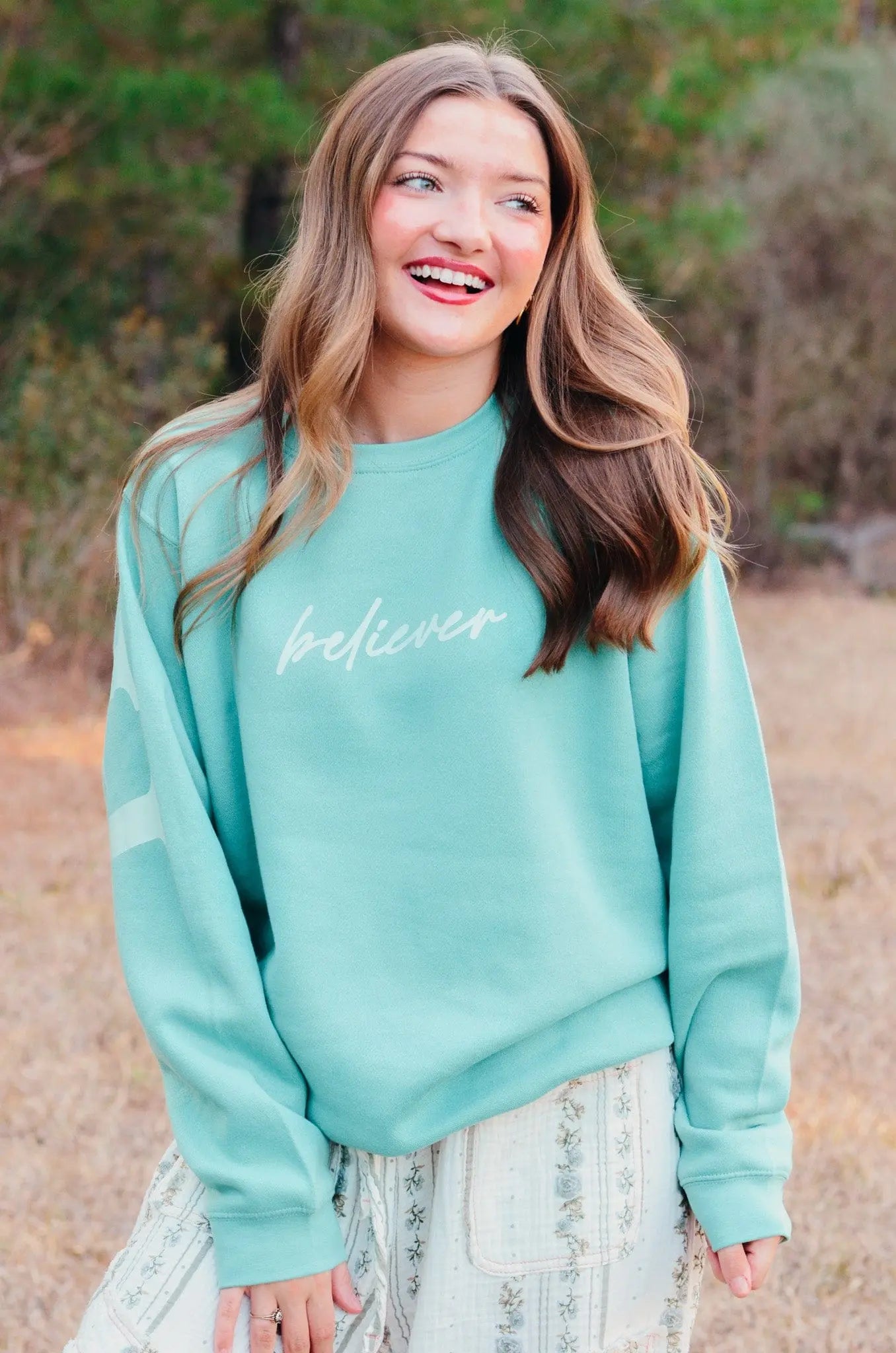 Woman in teal "believer" sweatshirt from Main Street Collective smiles outdoors.