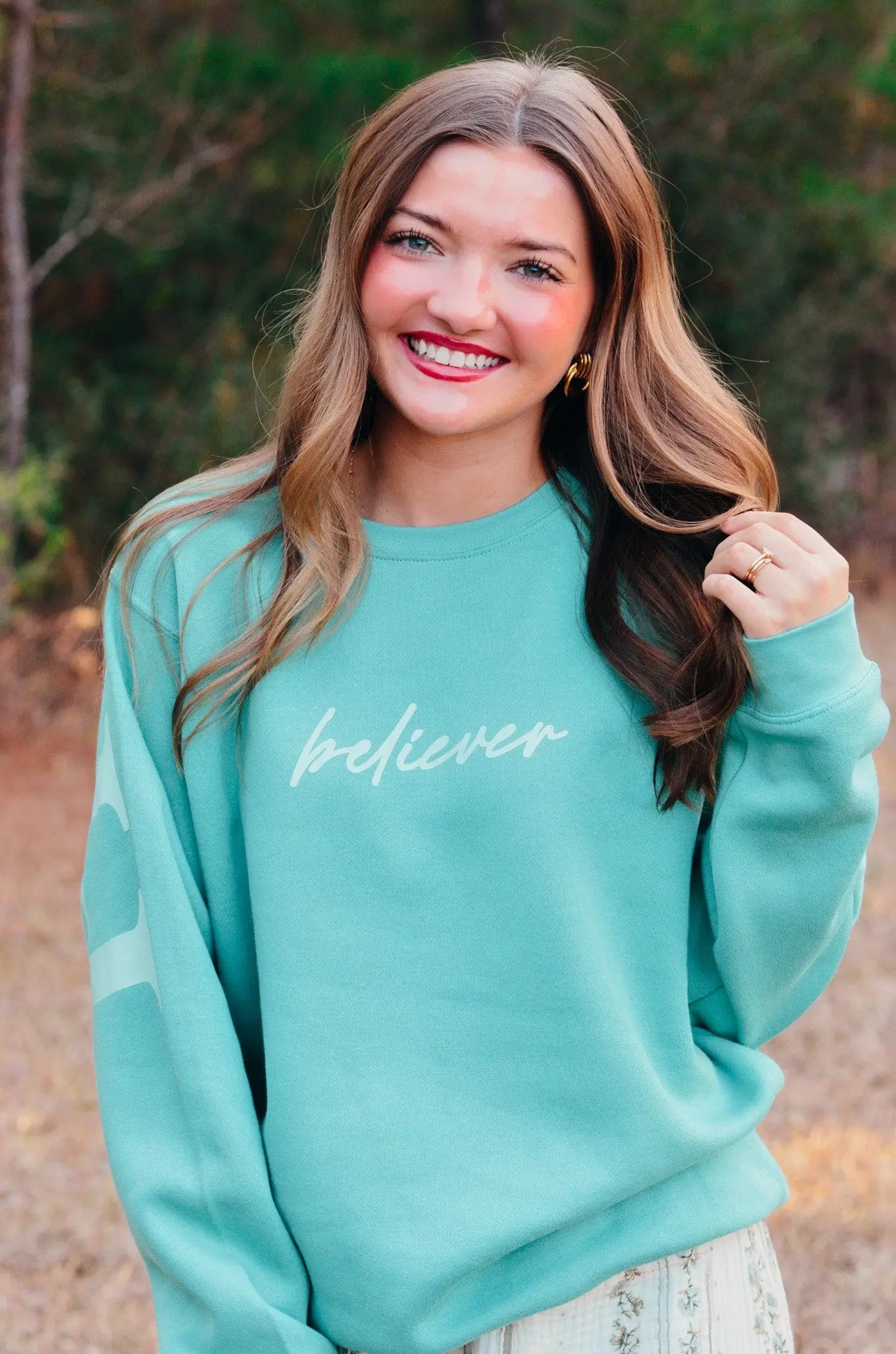 Woman in teal "believer" sweatshirt with long wavy hair, smiling outdoors in a field.