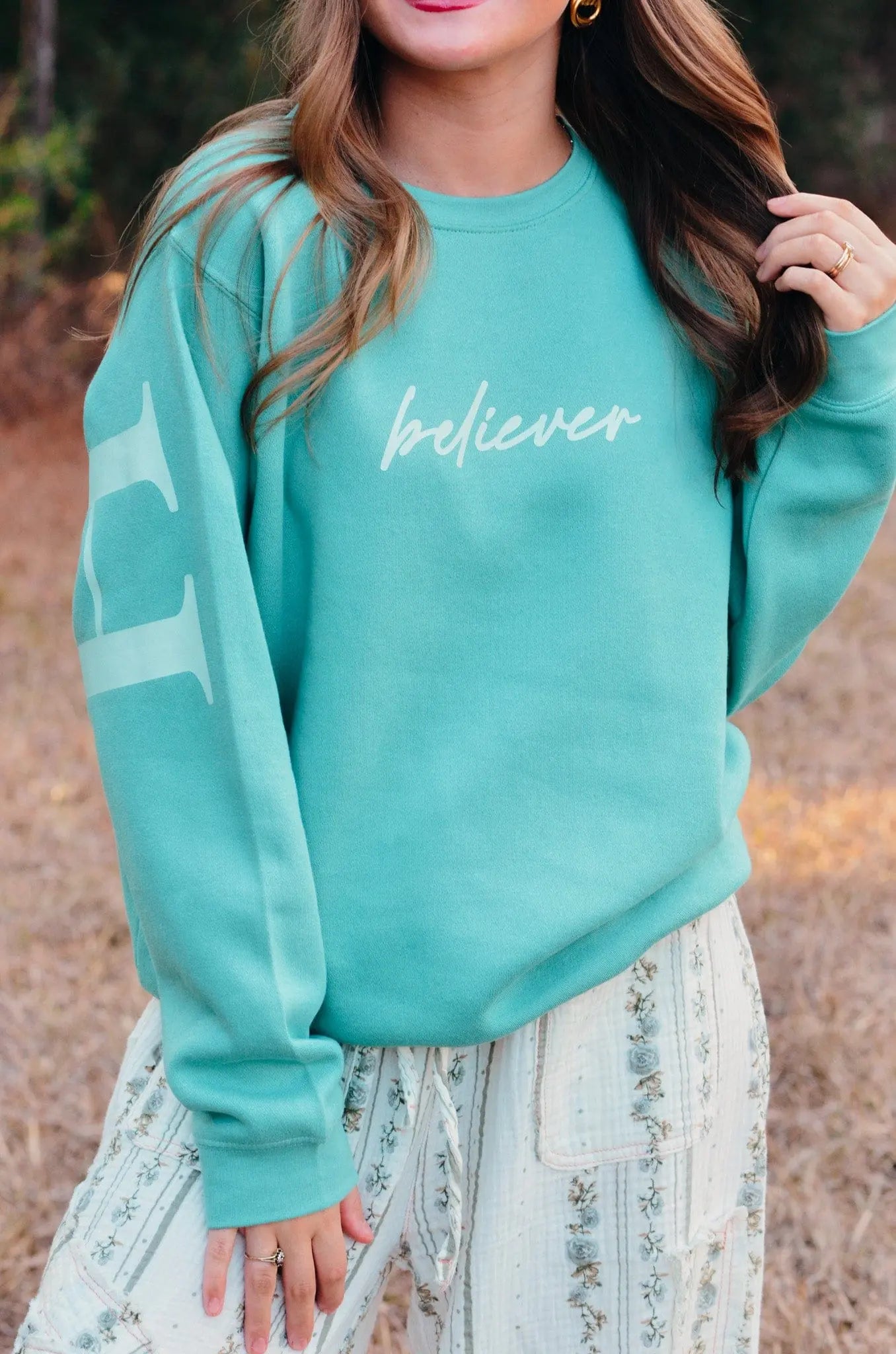 Teal sweatshirt with 'believer' print & stripe on sleeve. Model wears floral pants; outdoor setting. Casual style. Long sleeves, crew neck.