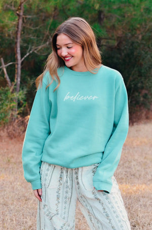 Woman in a teal "believer" sweatshirt and floral pants stands outside.