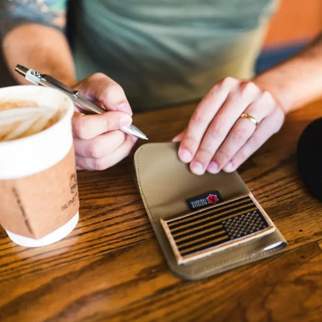 Man writing in a notebook with a coffee cup and a tan wallet featuring an American flag patch.