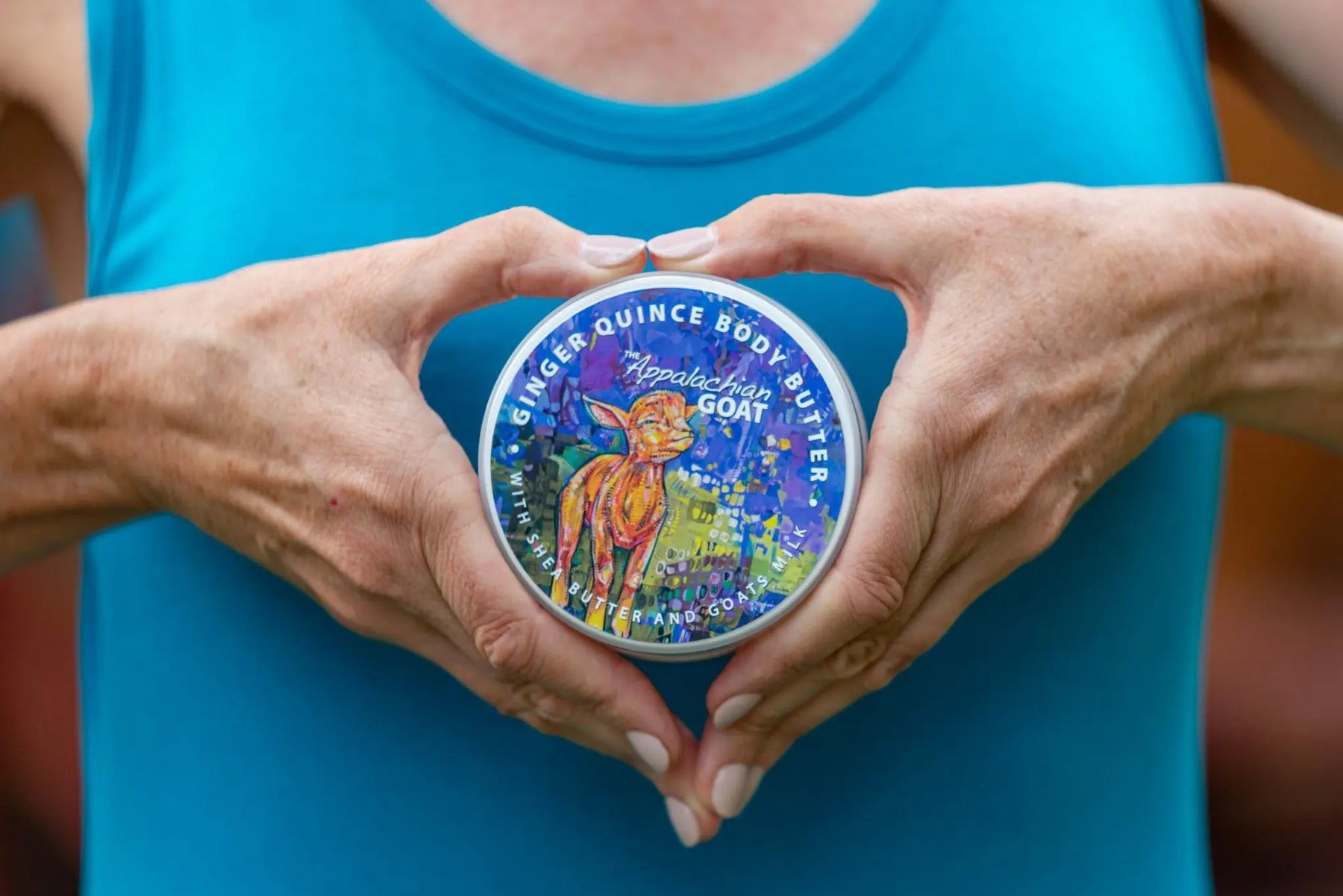 Hands forming a heart shape around a round tin of Ginger Quince Body Butter with goat illustration.
