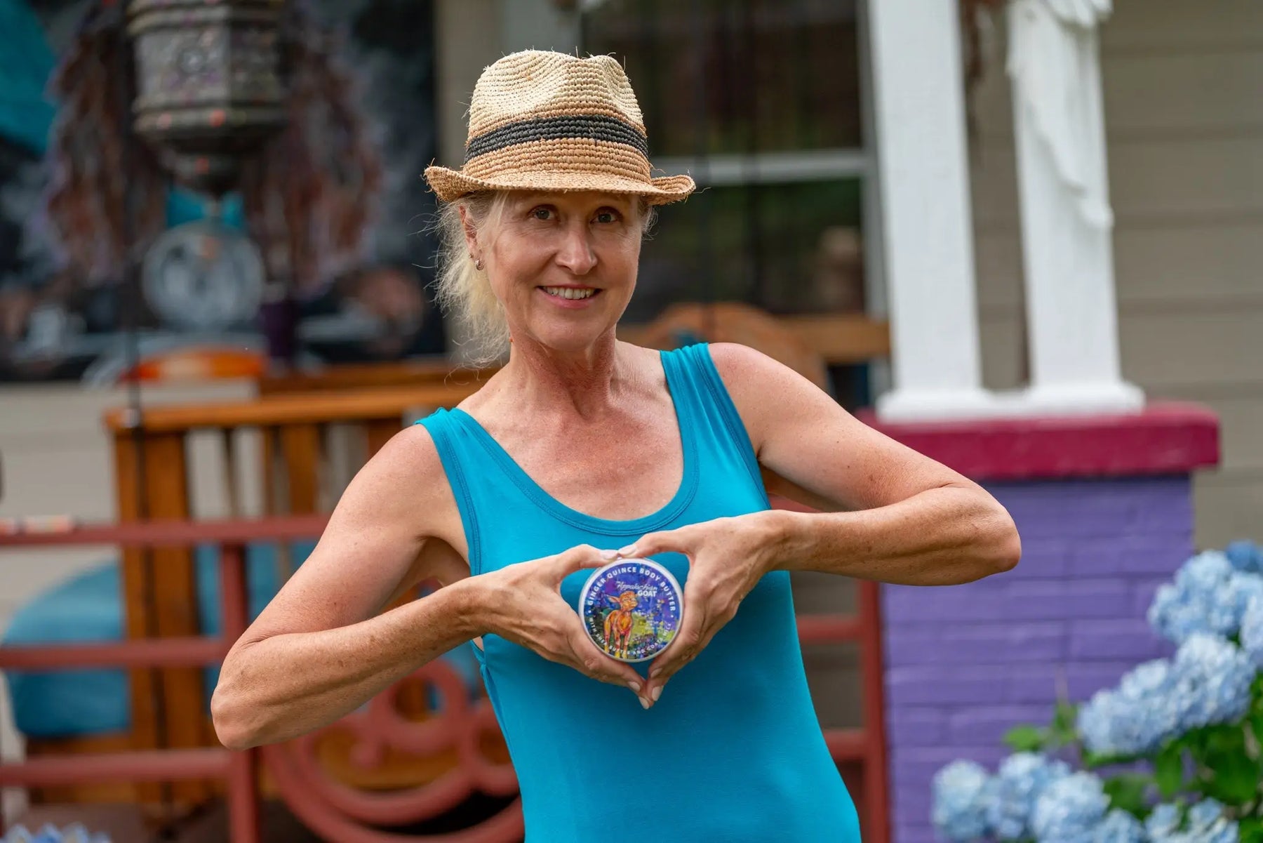 Woman in blue tank top and straw hat forms heart shape with hands around Ginger Quince Goats Milk Body Butter.