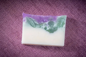 A slice of goat milk soap with purple, green, and white layers on a purple textured surface.