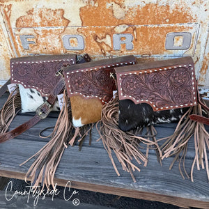 Tooled leather purse with cowhide front and leather fringe, displayed on a wooden surface.