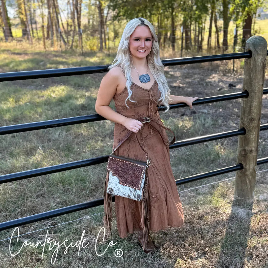 Woman wearing a brown dress holding a tooled leather and cowhide purse with fringe.