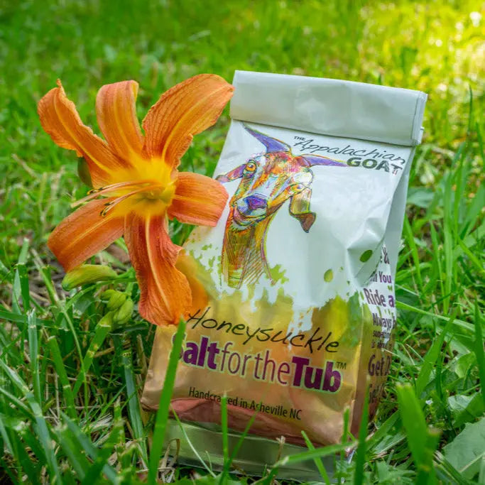 Honeysuckle Goat Milk Bath Salts next to an orange flower on grass.