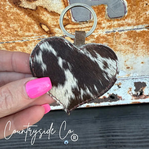 Brown and white cowhide heart-shaped keychain with leather backing held by a hand.