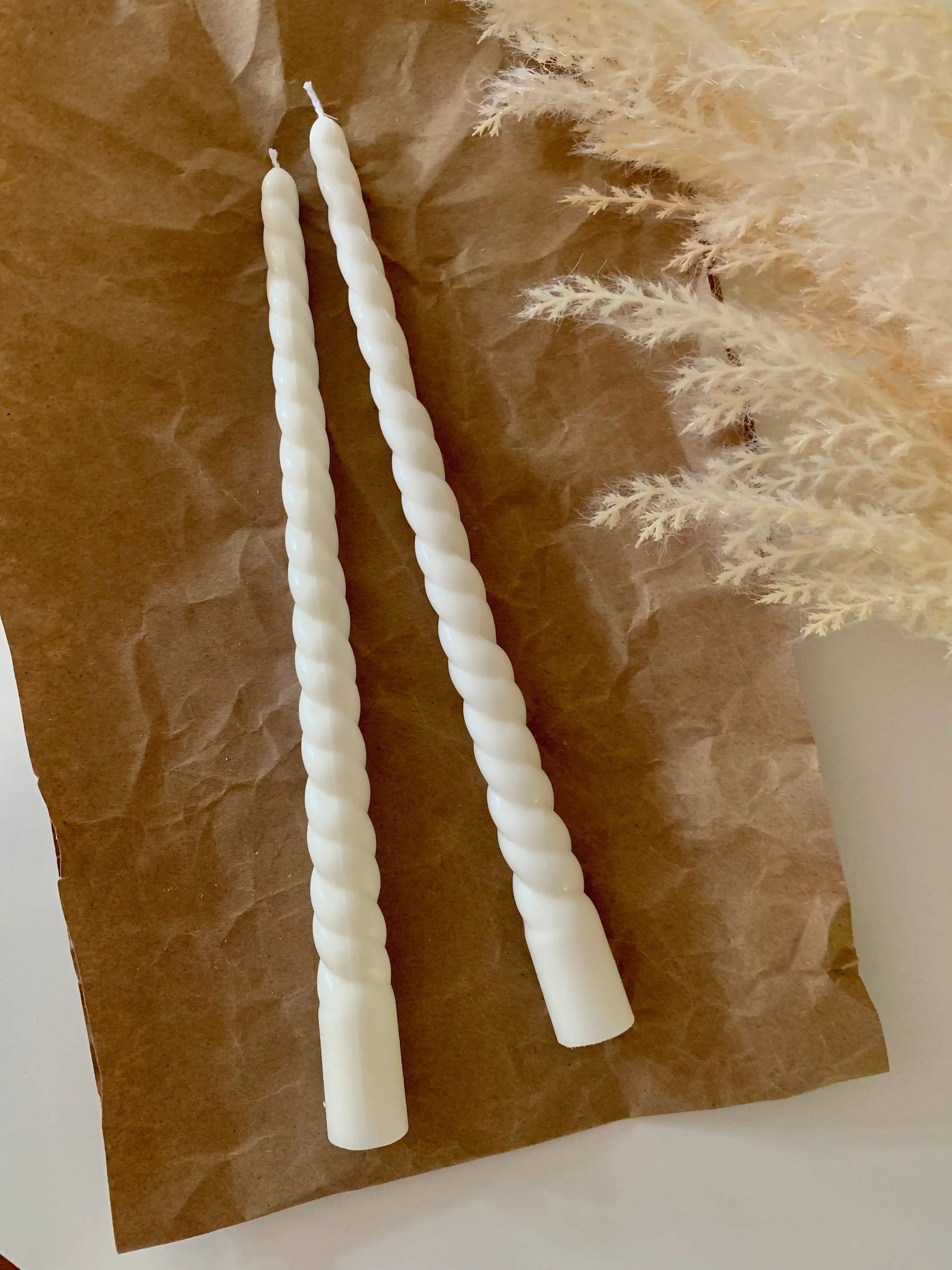 Two white twisted taper candles on brown crumpled paper with dried plants.