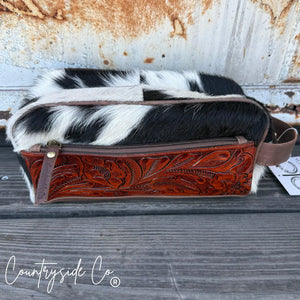Tooled leather and cowhide toiletry bag with zipper closure on wooden surface.