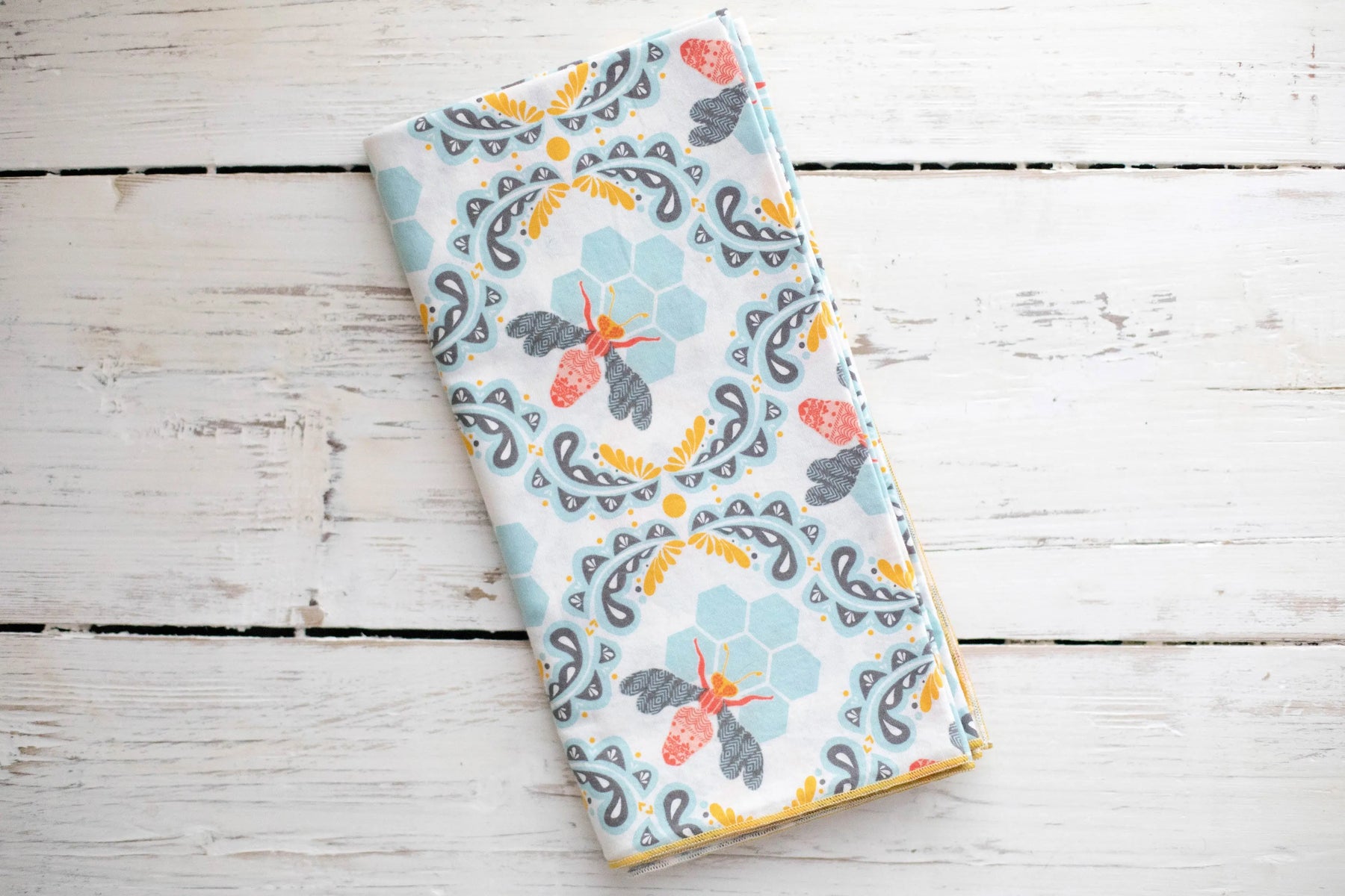 White cloth napkins with a repeating pattern of bees, hexagons, and floral designs in blue, orange, and gray.