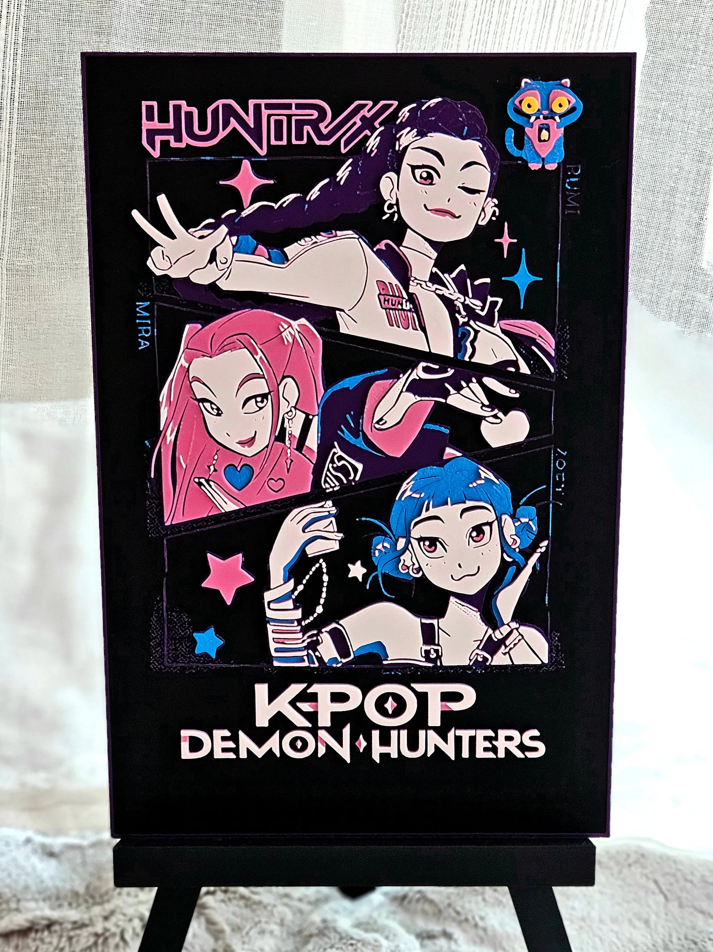 A K-Pop Demon Hunters poster featuring anime-style characters in pink, blue, and black on a stand.