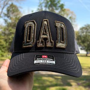 Black trucker hat with embroidered camouflage 'DAD' letters and Richardson tag.