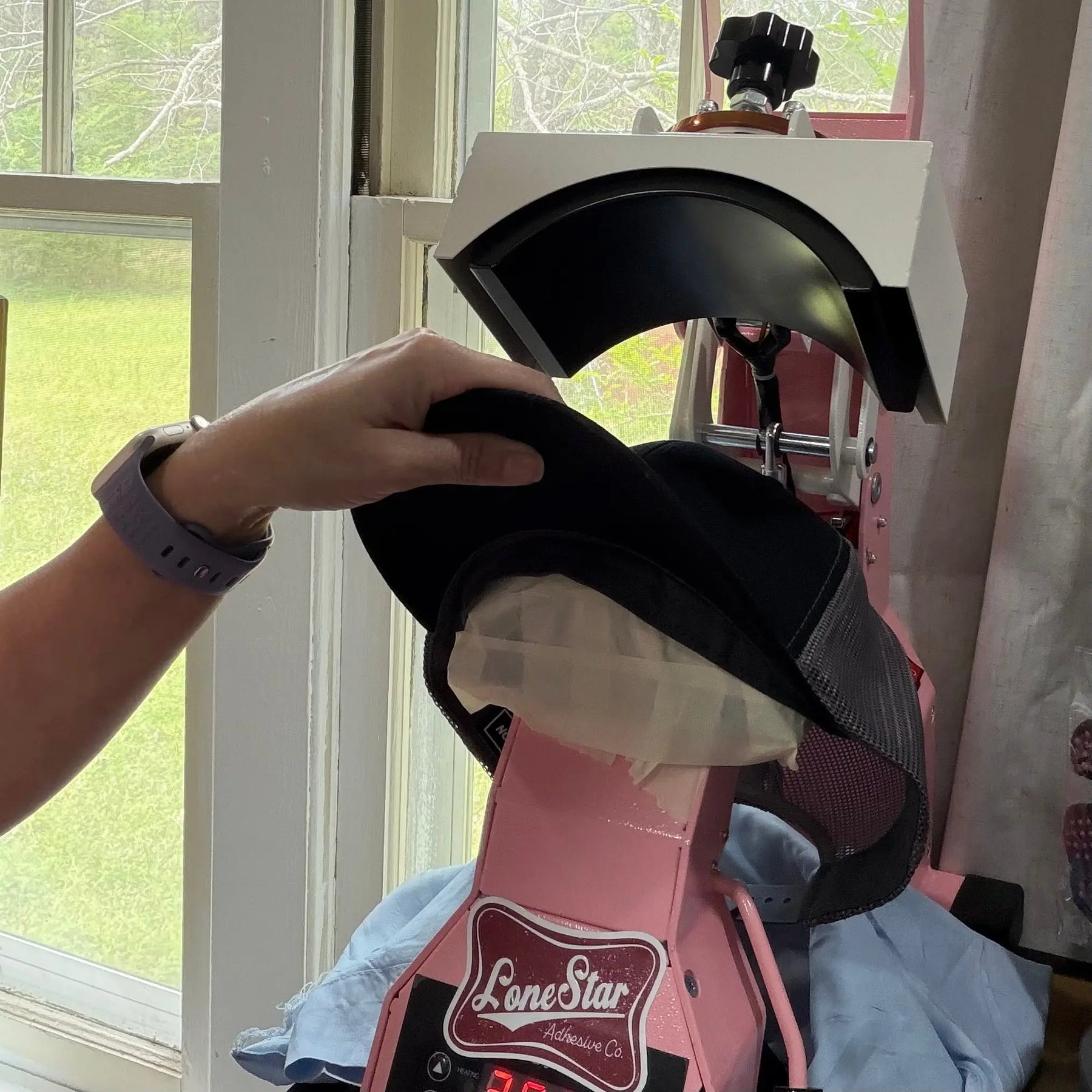 Hand placing a custom appliqué letter hat onto a pink heat press machine.