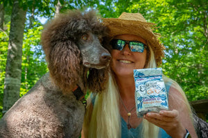 Woman in straw hat holds lavender and arnica bath salts bag next to a poodle.