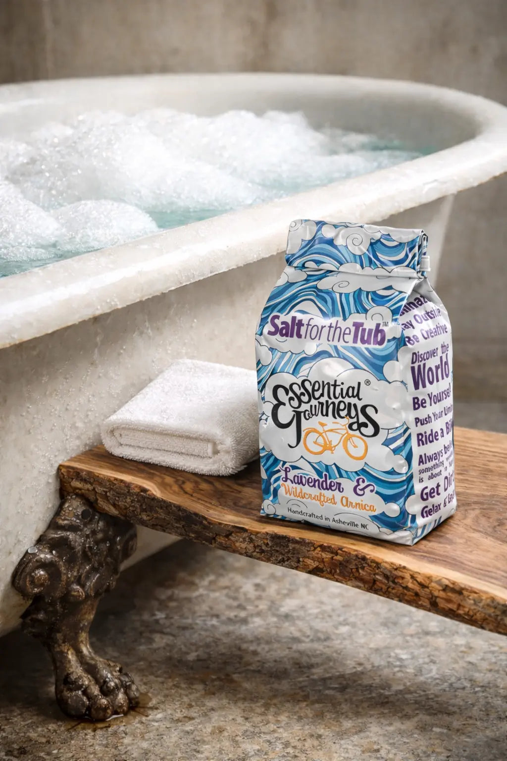 Essential Journeys Lavender & Arnica bath salts with foamy bathtub in background.