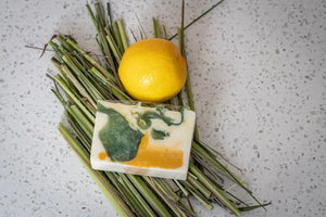 Bar of lemongrass goat milk soap slice with green and yellow swirls, next to a lemon and grass on a speckled surface.