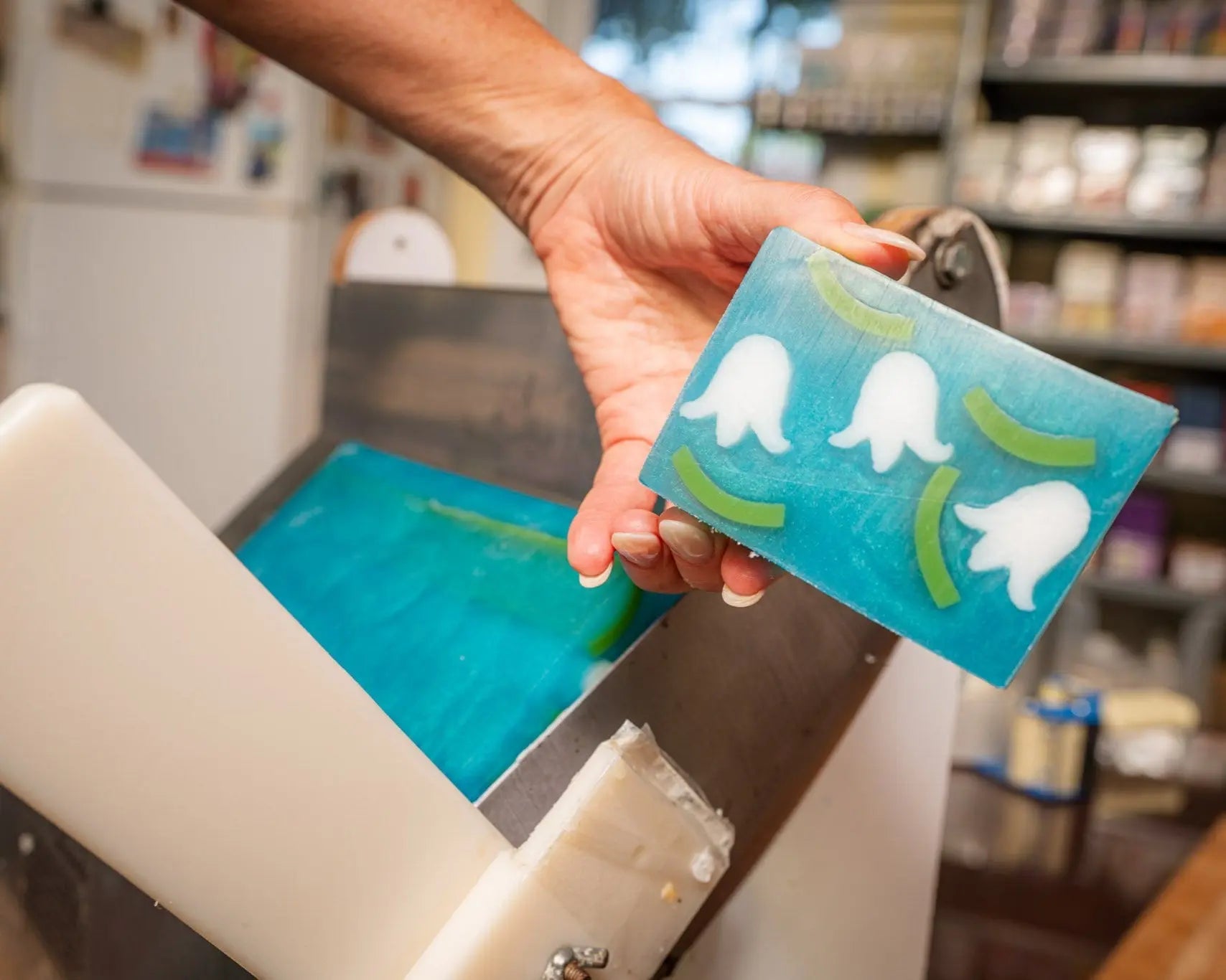 A hand holds a blue soap slice with white lily of the valley designs and green accents.