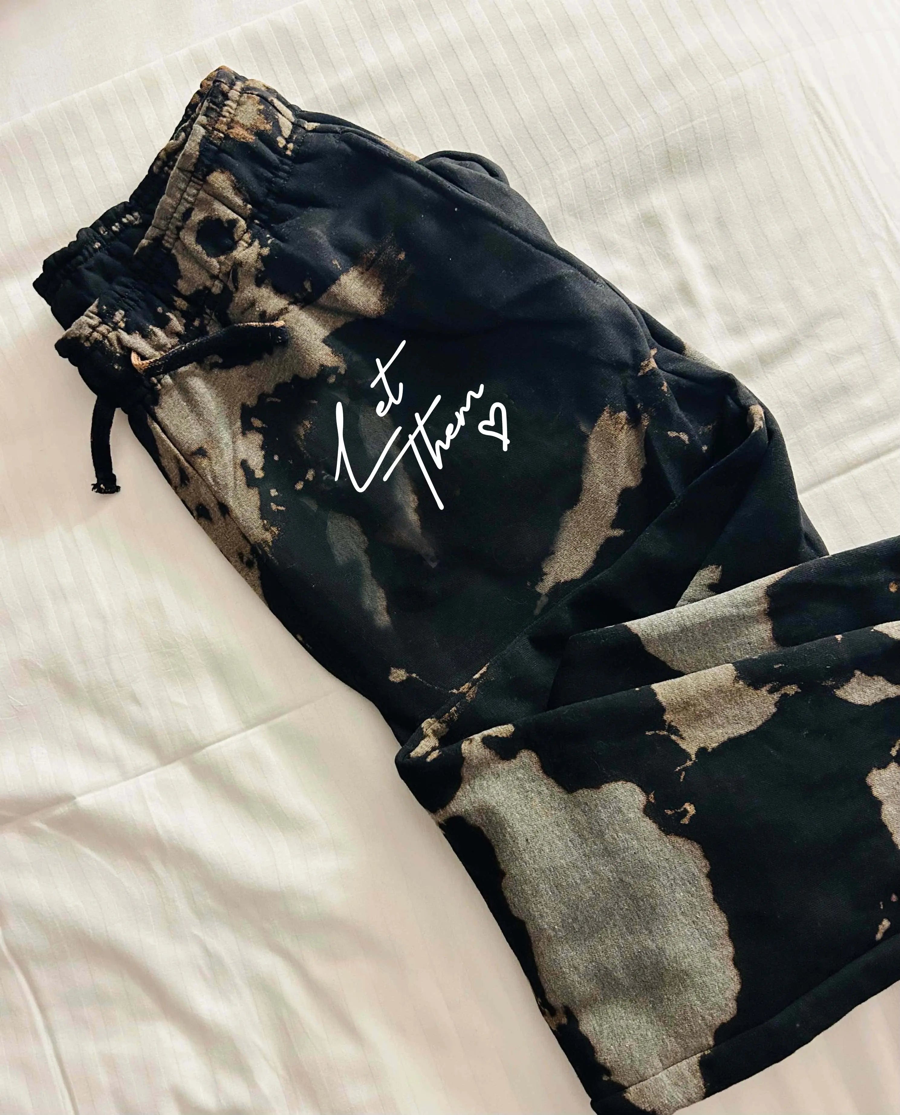Black and gray tie-dye joggers with drawstring and "Let Them" graphic on a white bedsheet.