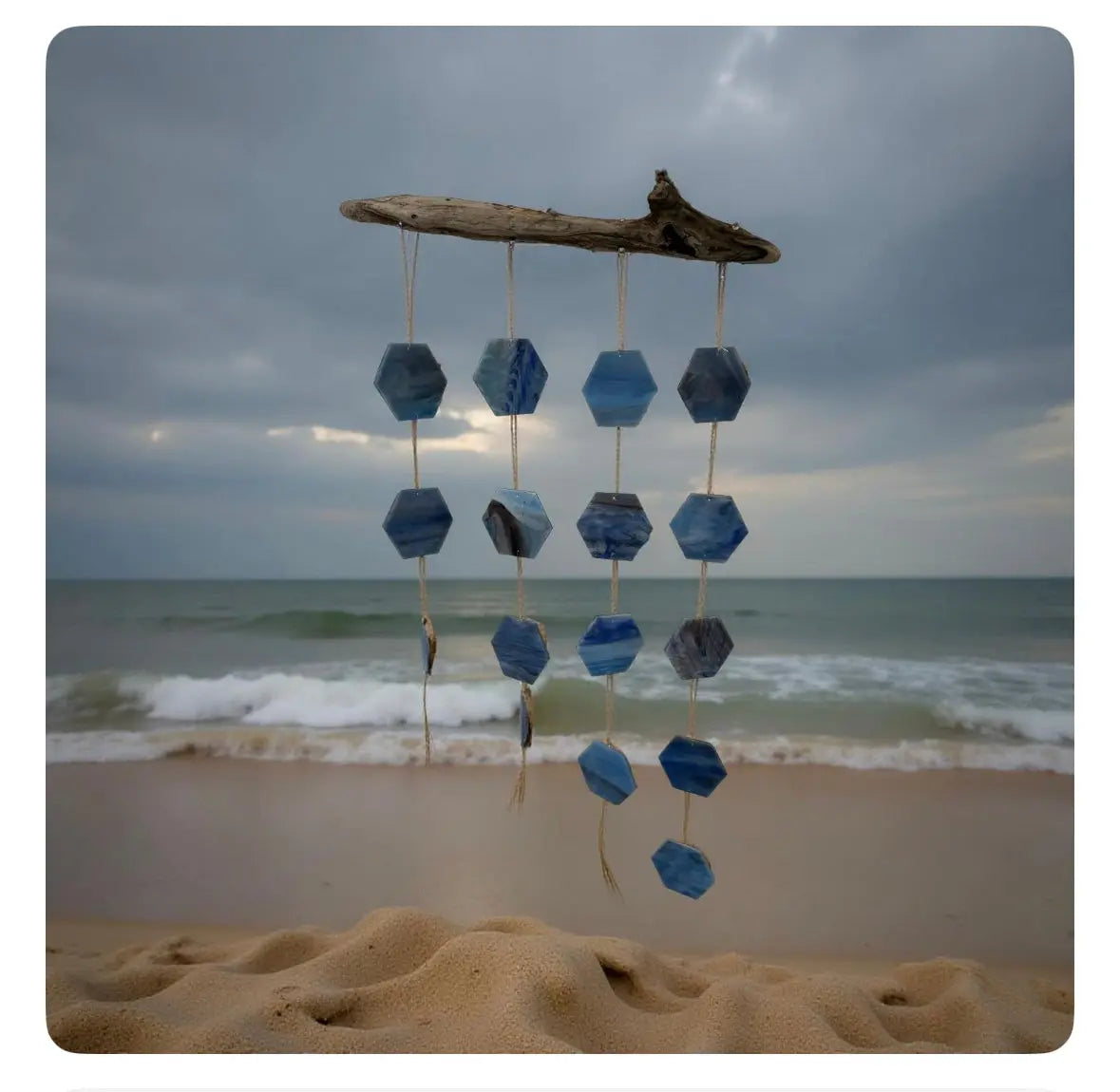 Driftwood wind chime with blue mosaic glass hexagons hanging in front of a beach.