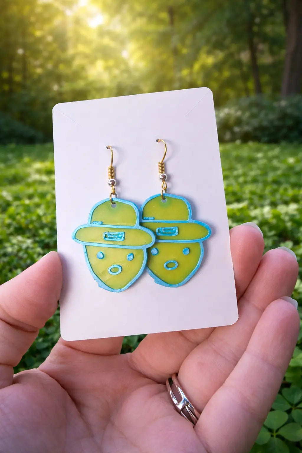 Leprechaun earrings: A pair of handmade green and blue leprechaun earrings with gold hooks on a white card in a hand.