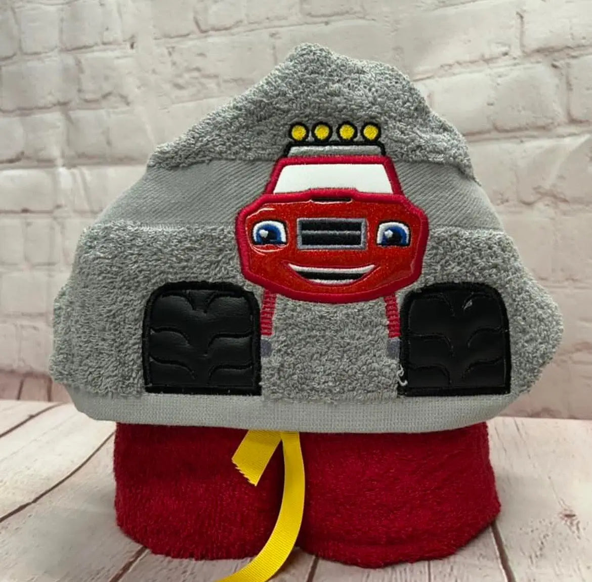 Monster truck hooded towel for kids: A gray towel forms the hood with a red truck design and black tires. Yellow ribbon ties the base.
