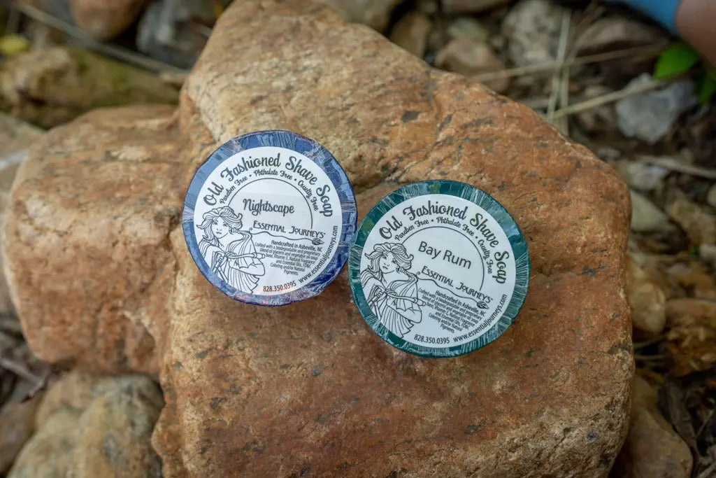 Two round Essential Journeys Old Fashioned Shave Soaps, Nightscape and Bay Rum scents, on rocks.