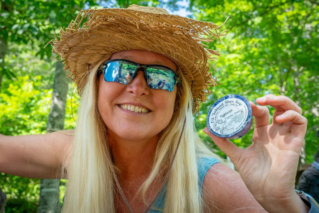 Woman wearing a straw hat and sunglasses holds a round tin of "Old Fashioned Shave Soap Nightscape"