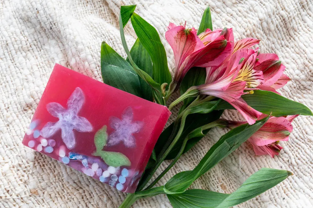 Pink handmade soap with floral design next to pink alstroemeria flowers on a textured cloth.