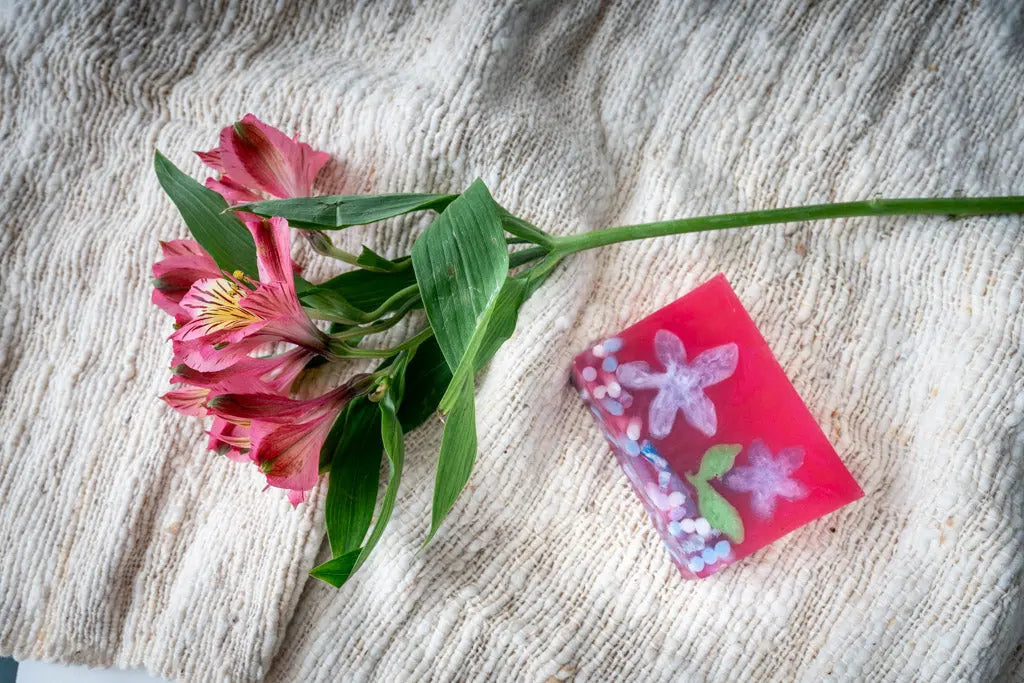 Pink soap slice with painted flowers next to pink alstroemeria flowers on textured fabric.