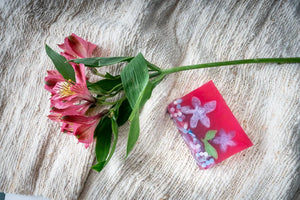 Pink soap slice with painted flowers next to pink alstroemeria flowers on textured fabric.