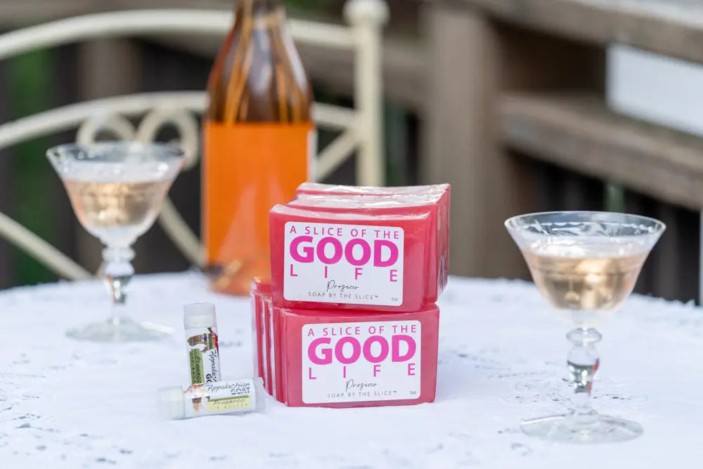 Stack of pink Prosecco soap bars with cocktail glasses and lip balms on a white tablecloth.