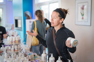 Woman presenting ROSEMARY MINT ARNICA SALVE 8 oz. Essential Journeys product at a table.
