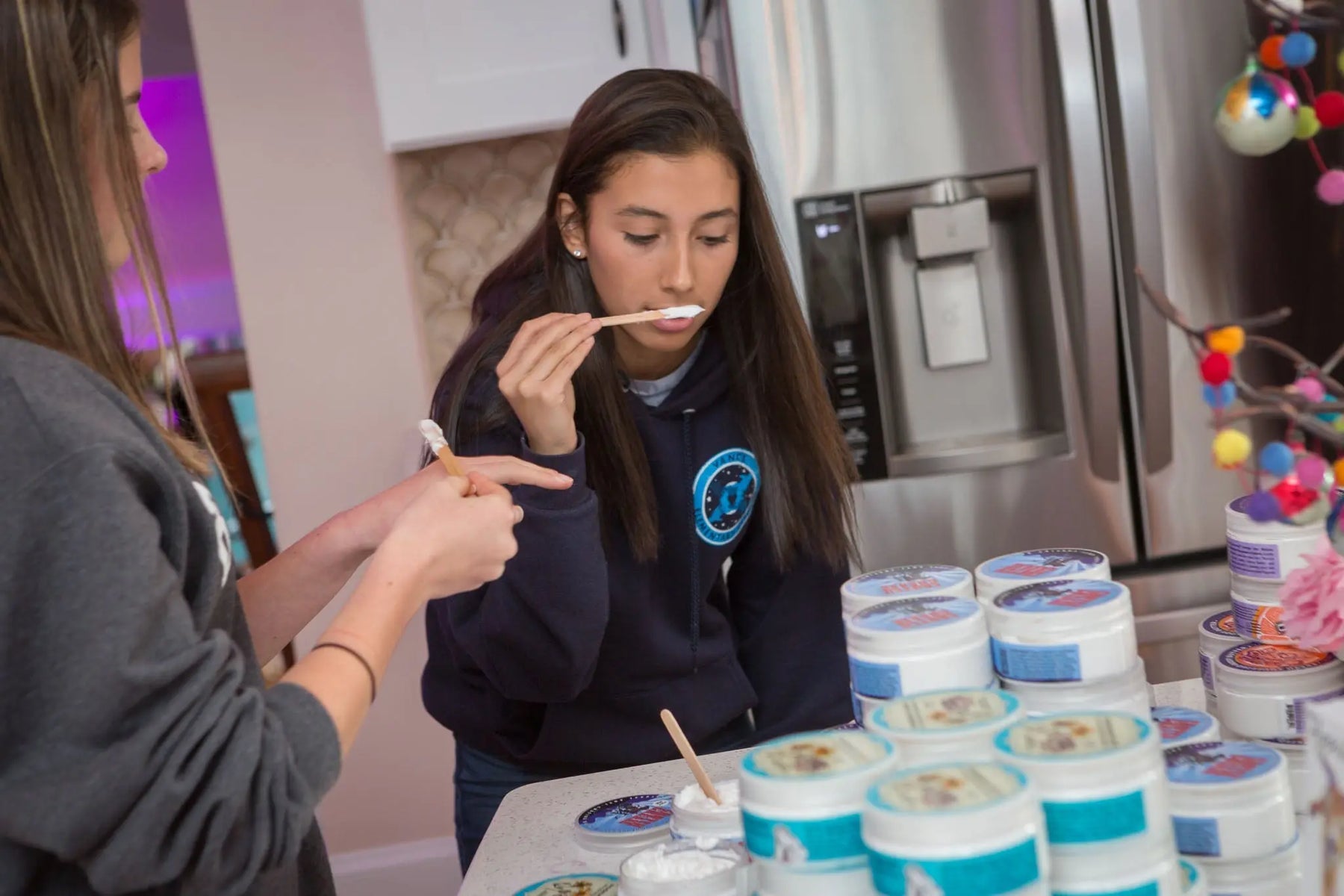 Two young women sampling Rosemary Mint Arnica Salve from jars with wooden applicators.