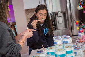 Two young women sampling Rosemary Mint Arnica Salve from jars with wooden applicators.