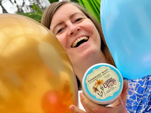 Woman holding a Rosemary Mint Arnica Salve jar with balloons and greenery in the background.