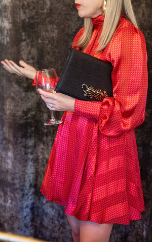 Woman in red dress holding a black clutch with gold leaf detail and a wine glass.