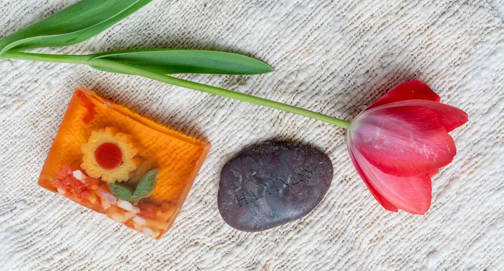 Orange sunflower soap slice with red tulip and 'happiness' stone on textured fabric.