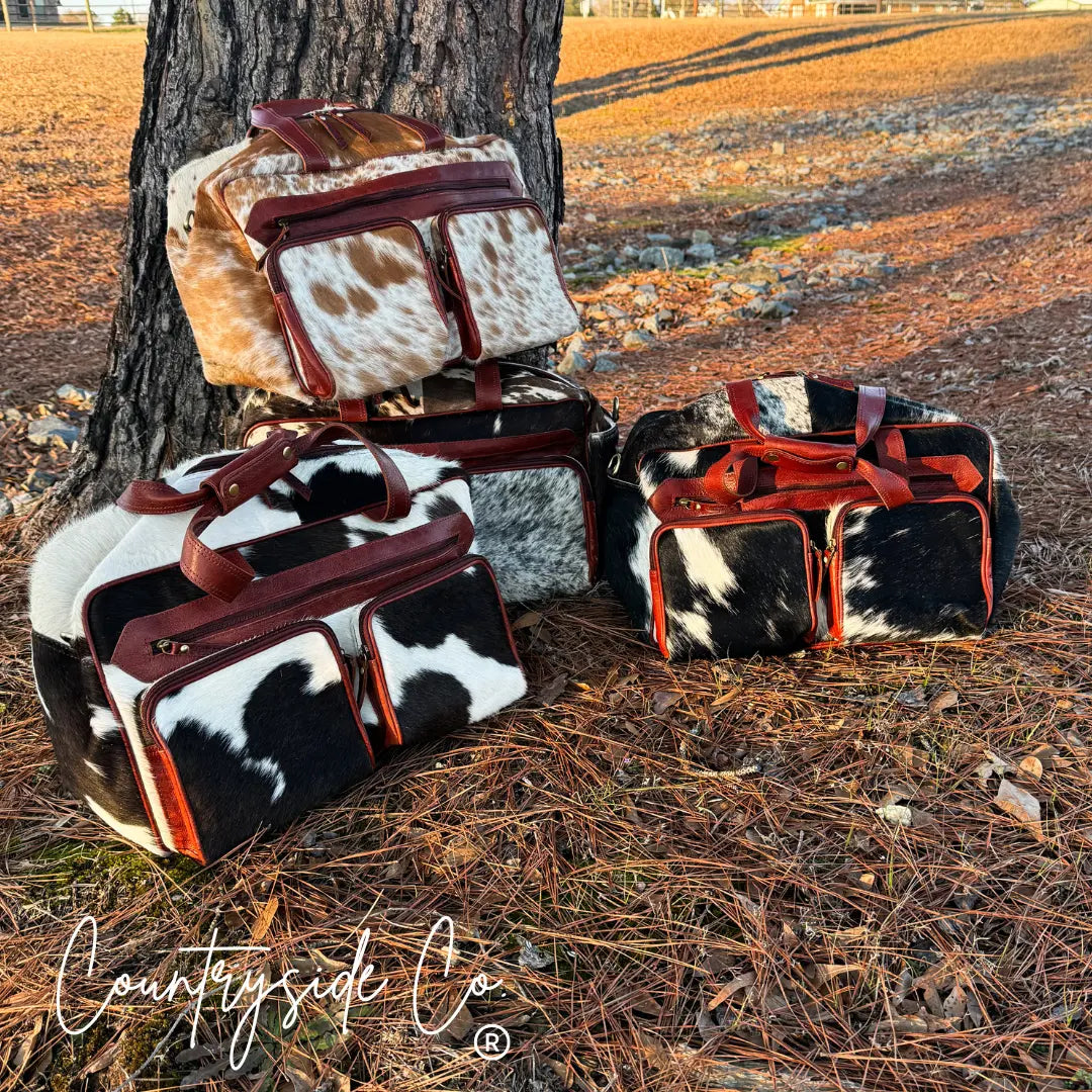 Multiple cowhide and leather duffle bags arranged by a tree outdoors.
