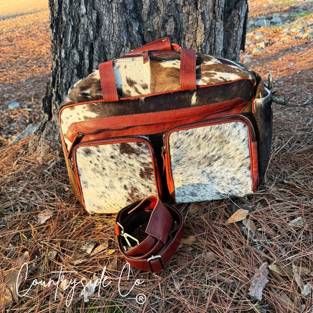 Rustic cowhide duffle travel bag with rust-red leather trim and shoulder strap.