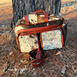 Rustic cowhide duffle travel bag with rust-red leather trim and shoulder strap.