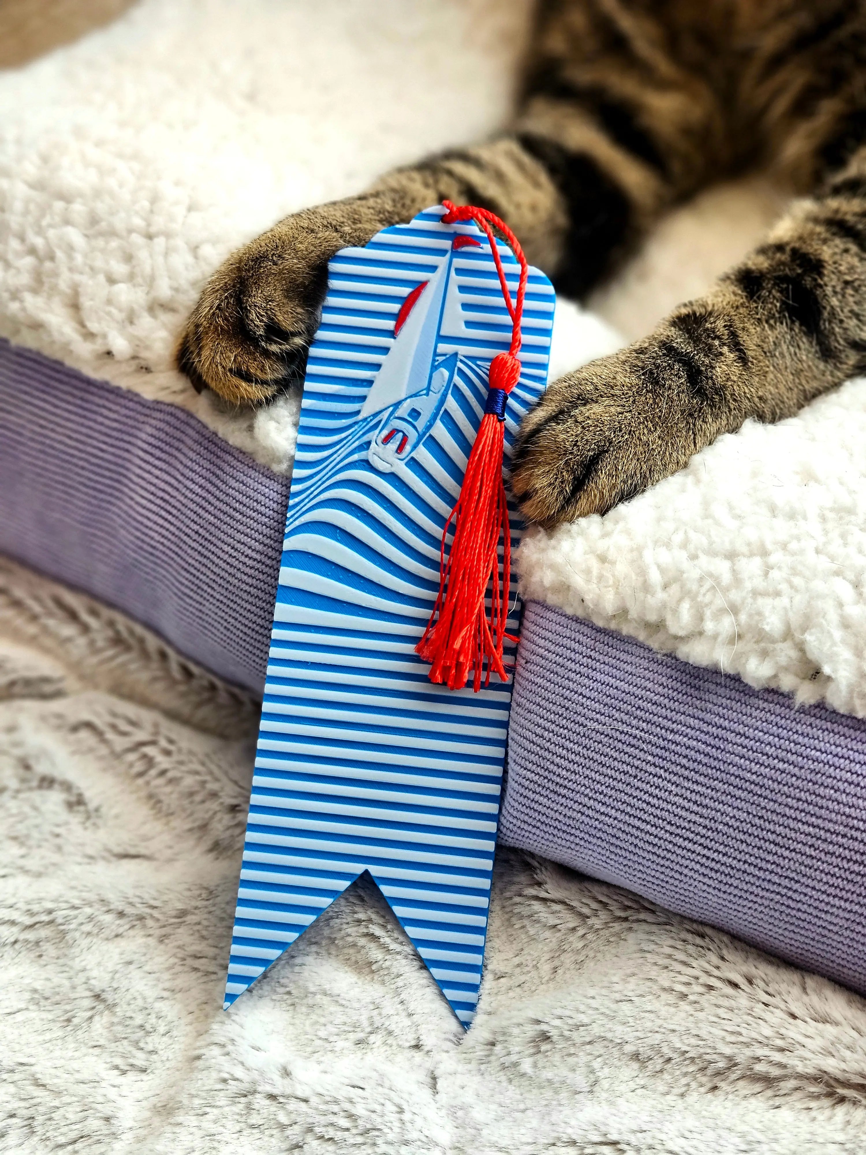 Nautical sailboat bookmark with red tassel held by tabby cat paws on a soft bed.