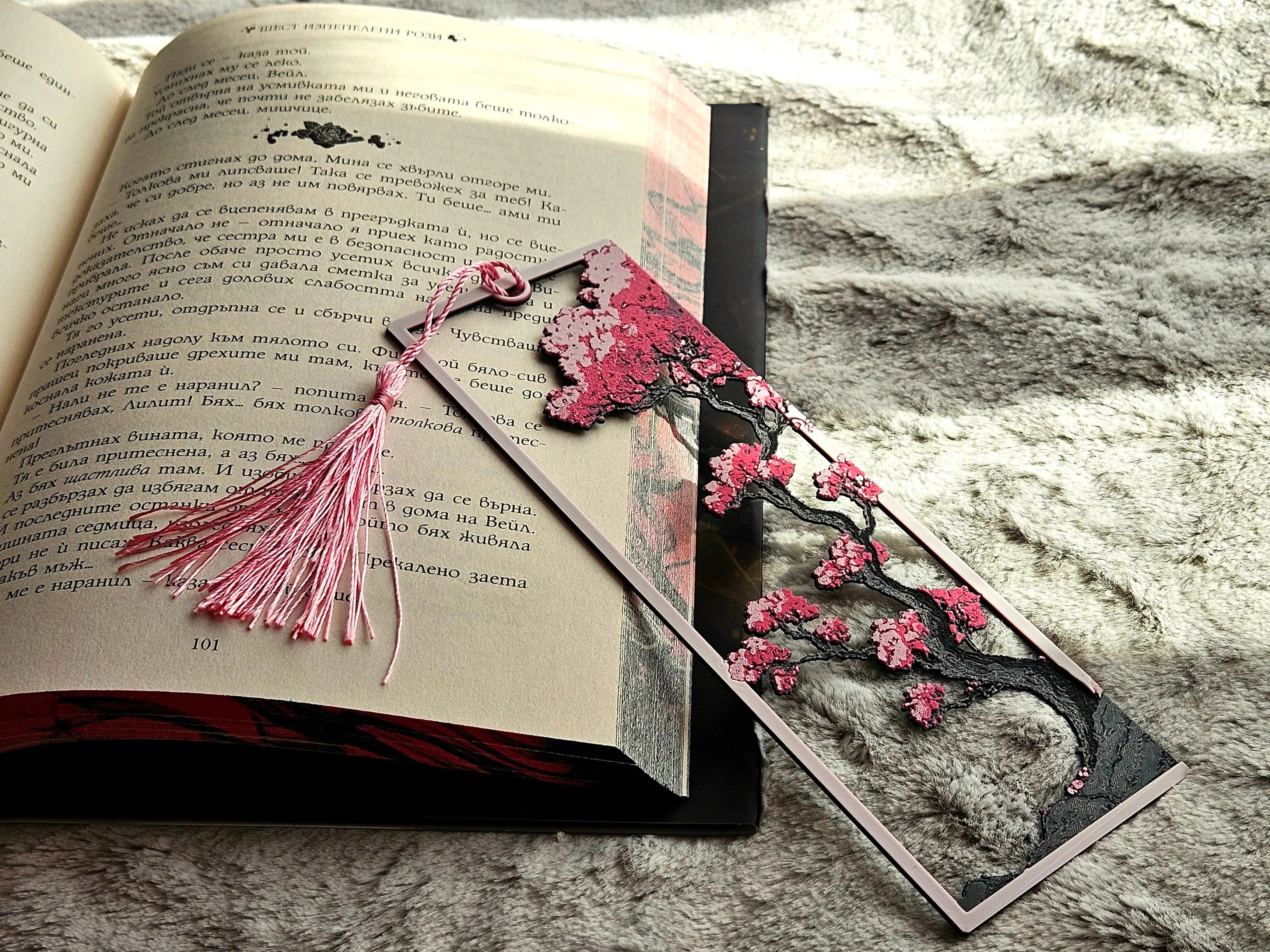 Cherry blossom samurai bookmark with tassel resting on open book. Pink, black, and beige colored rectangular framed bookmark.