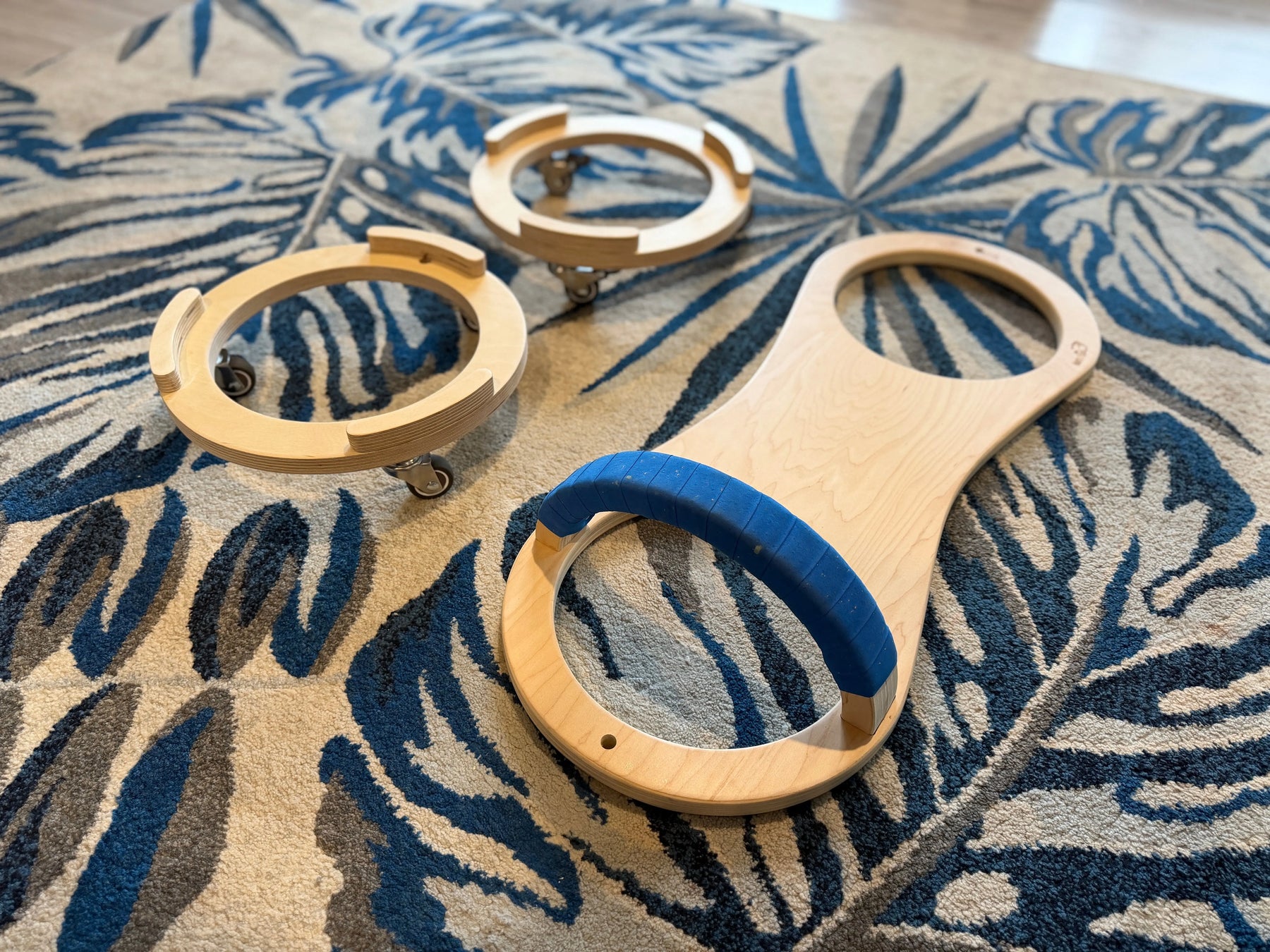 Wooden scooter board with blue handle and two rolling bases on a patterned carpet.