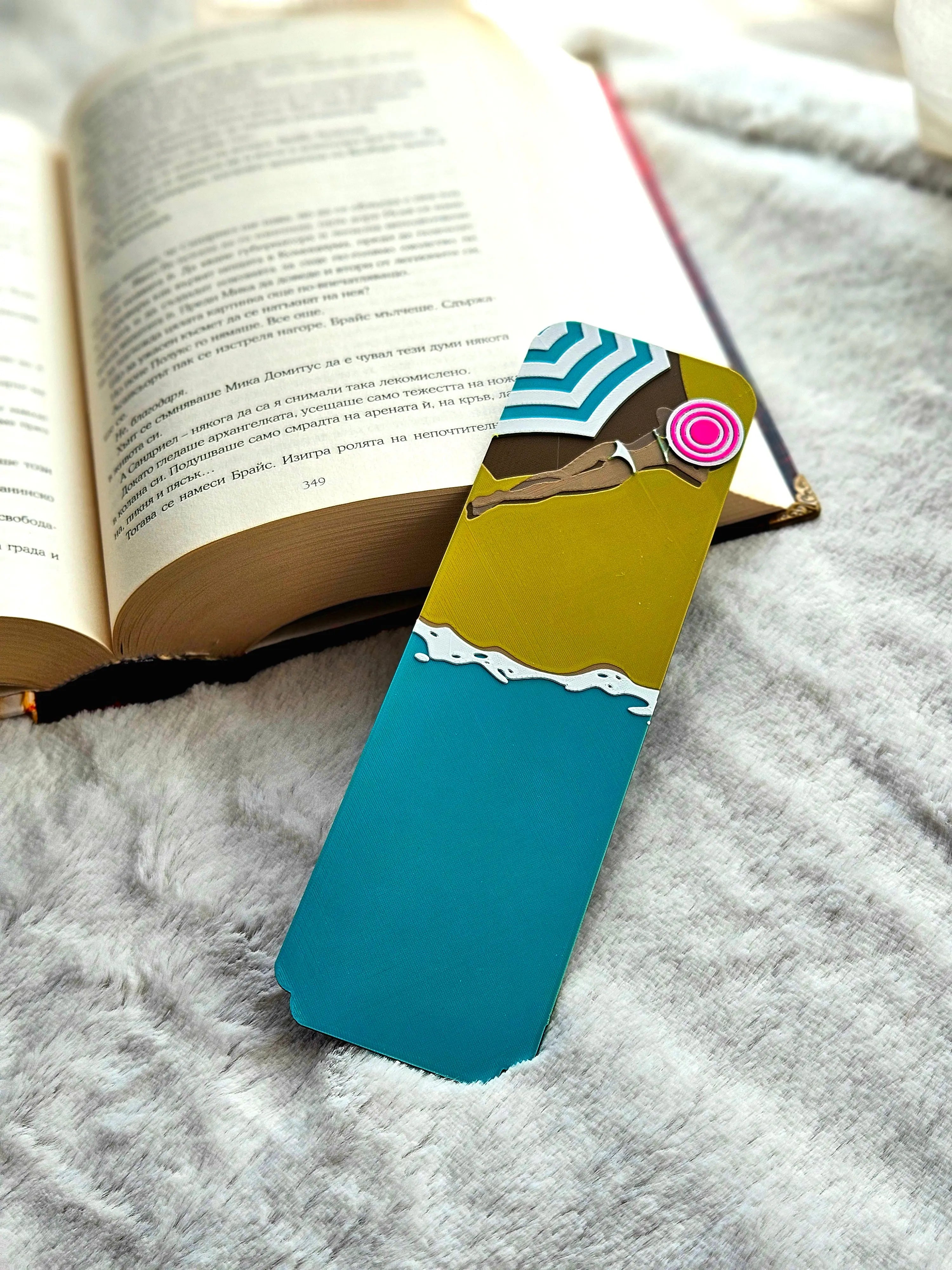 Bookmark with a beach scene showing an umbrella, sand, and ocean waves next to an open book.