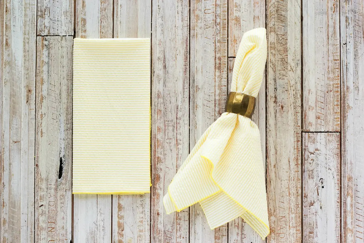 Yellow seersucker dinner napkins folded with a gold napkin ring on a wood background.