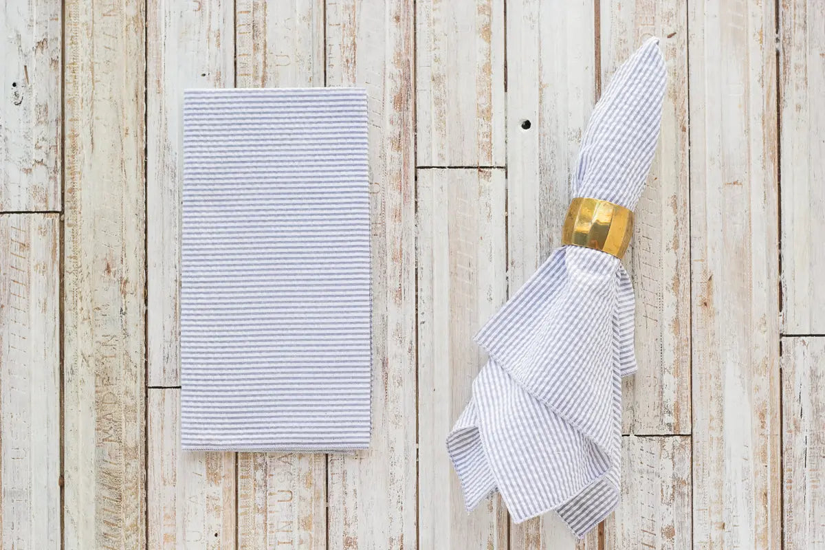 Seersucker dinner napkins with blue and white stripes and a gold napkin ring on a wood background.