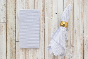 Seersucker dinner napkins with blue and white stripes and a gold napkin ring on a wood background.